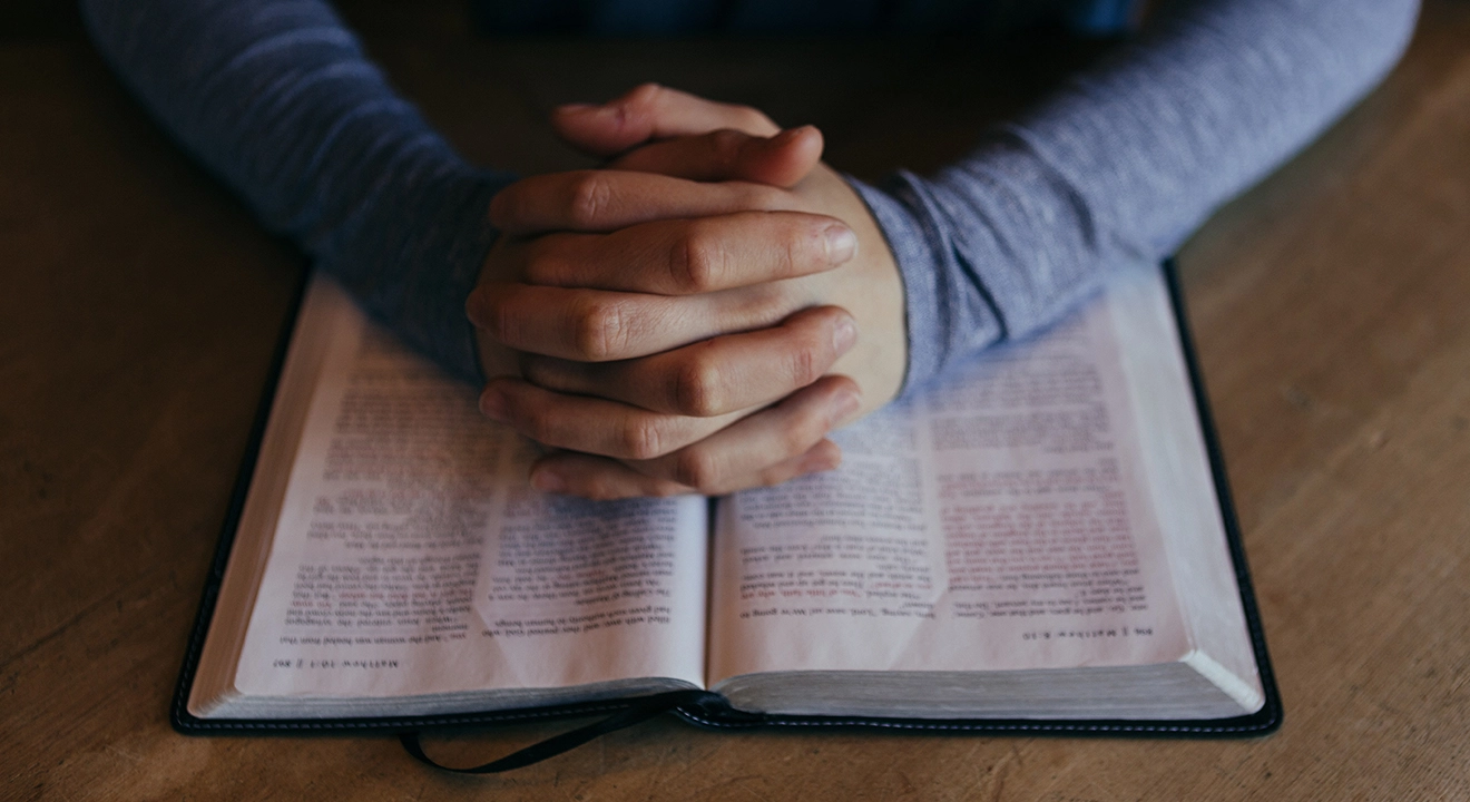 Folded hands resting on an open Bible on a desk